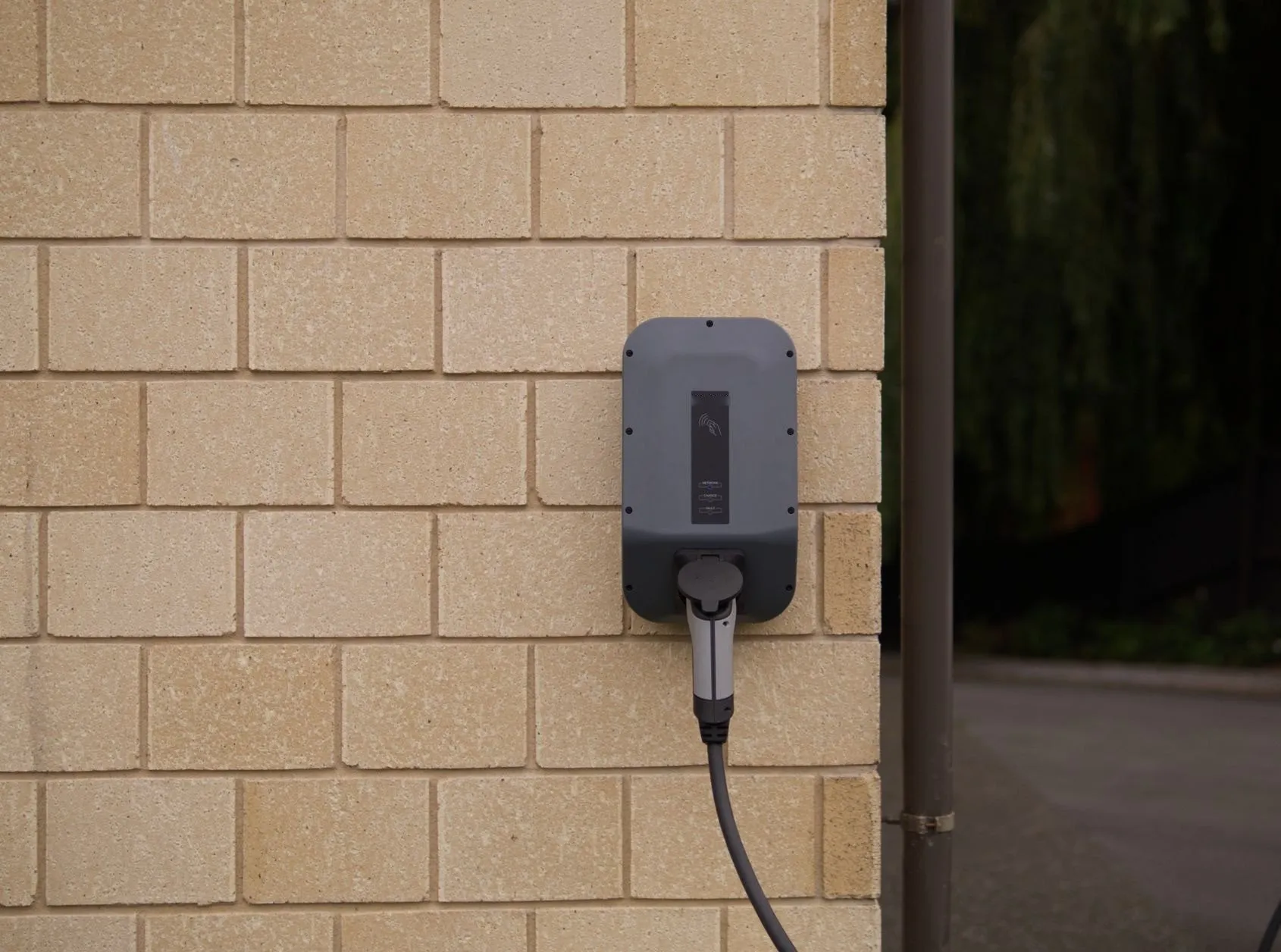 A grey home EV charger mounted on a pale brick wall with its cable plugged in