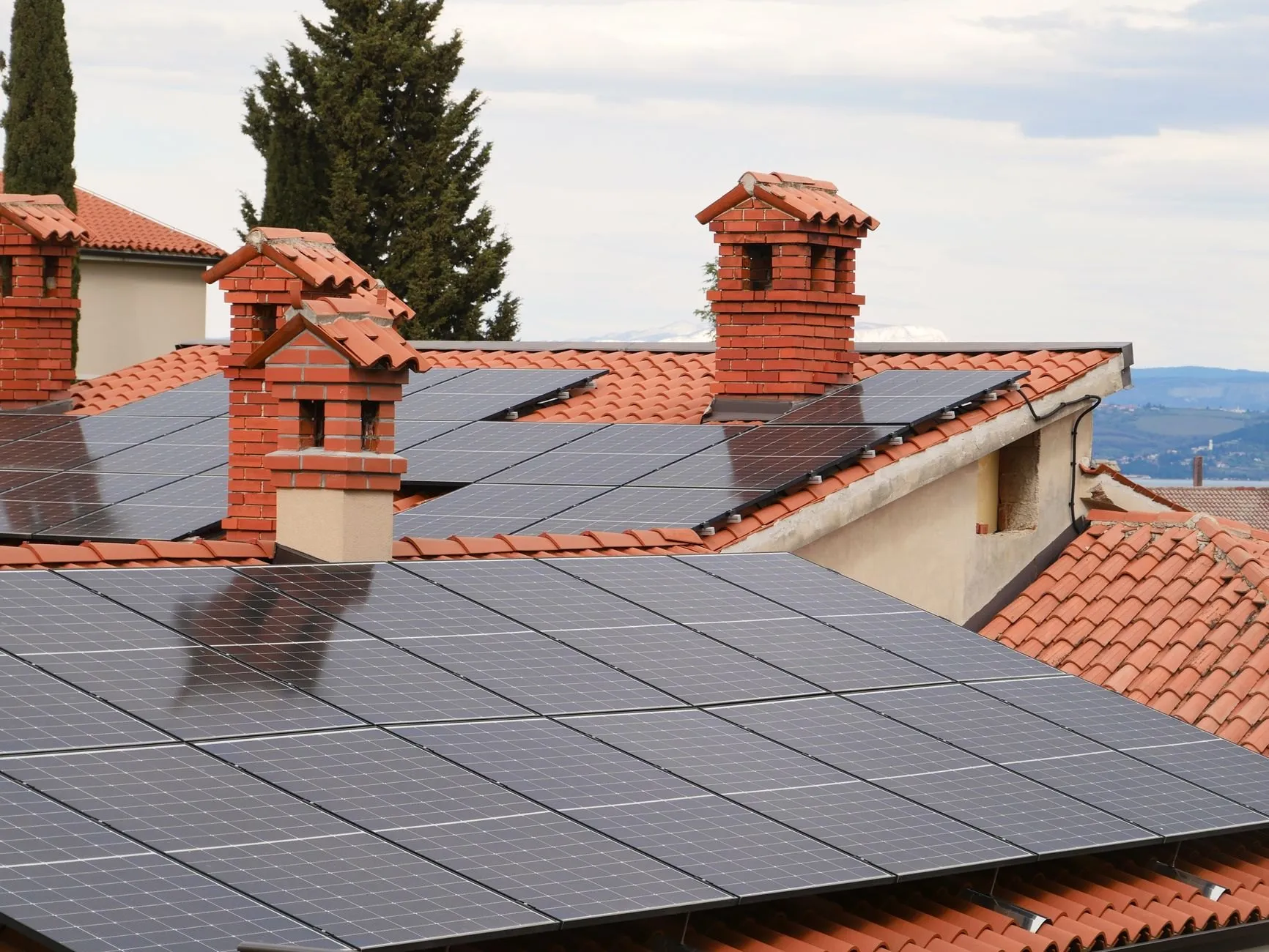 Solar panels on tiled roofs with brick chimneys under a cloudy sky